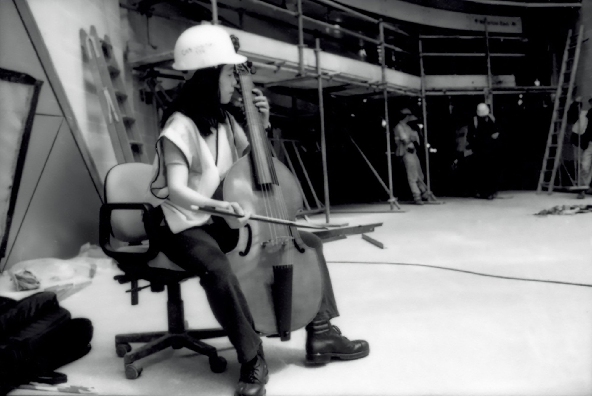 Image: Reiko Ichise wearing a hard-hat and high vis bib, plays the viola da gamba on an office chair on location during production of 13 Different Keys at the Atlantis Building, 1999. Photograph: Sarah Ainslie