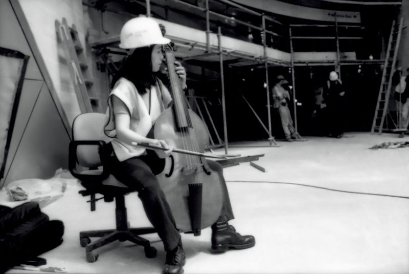 Image: Reiko Ichise wearing a hard-hat and high vis bib, plays the viola da gamba on an office chair on location during production of 13 Different Keys at the Atlantis Building, 1999. Photograph: Sarah Ainslie