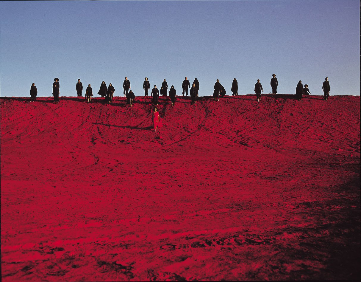 Image: Film still of Sussan Deyhim descending a red mountain, her followers appear on the horizon trailing down the slope behind her. Still: Shoja Azari, Sussan Deyhim, Ghasem Ebrahimian and Shirin Neshat