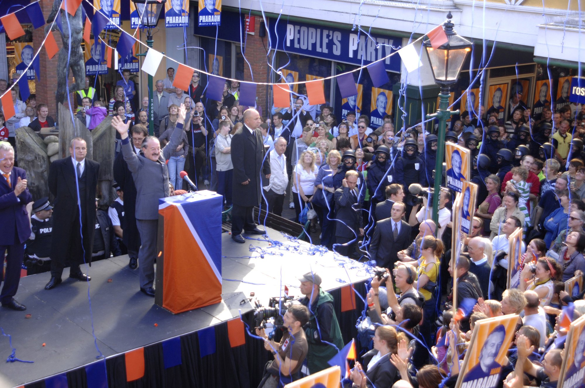A man wearing a suit stands on stage behind a lectern and waves at the crowd gathered around him. Film still from Exodus dir. Penny Woolcock shot in Margate 2006 