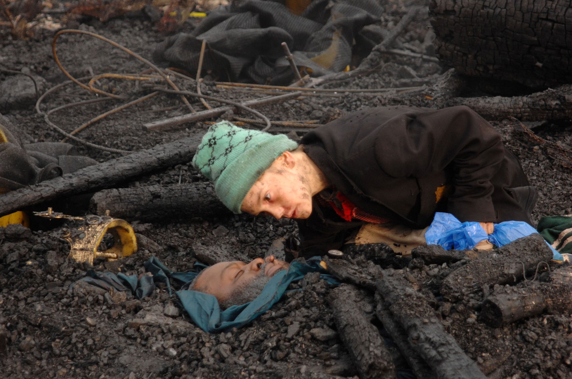 A young boy with a green woollen hat stares into the face of a man buried in charred earth. Film still from Exodus dir. Penny Woolcock.