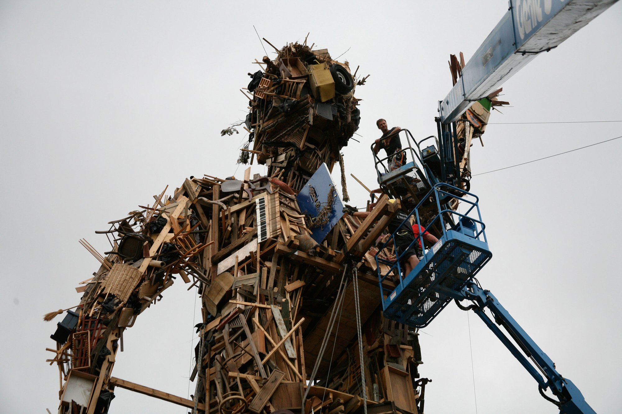 People on an elevated work platform add to an enormous effigy of a man, made from scrap wood. Photograph of Wasteman by Anthony Gormley which was created for the production of Exodus dir. Penny Woolcock in Margate 2006.