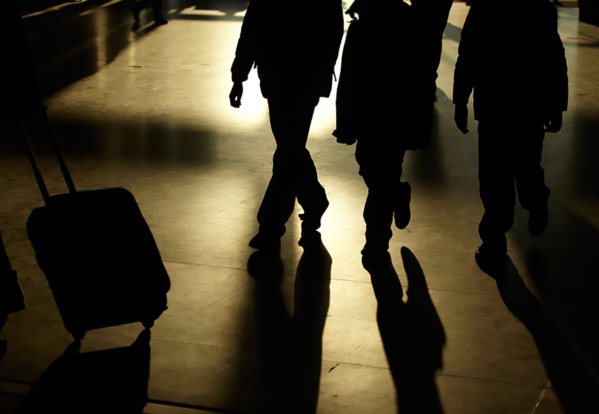 Image: Three silhouetted men walk across the concourse as another commuter pulls a wheeled suitcase out of shot. London St Pancras station 12 October 201. Photographer: Julian Abrams
