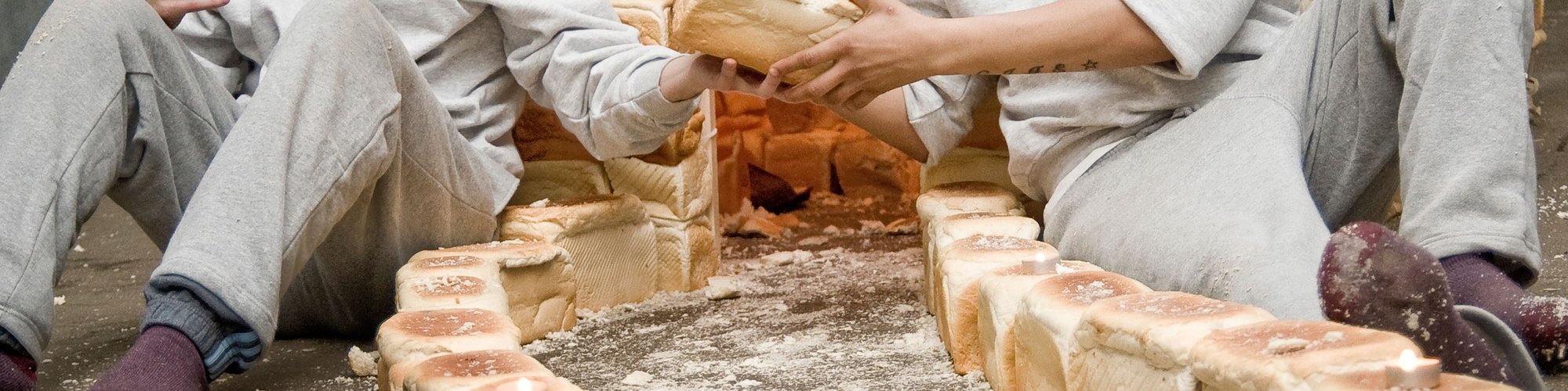 Image: Two actors sit either side of a road built out of white loaves. Behind them is a small structure built out of while loaves of bread. Photograph: Stephen King