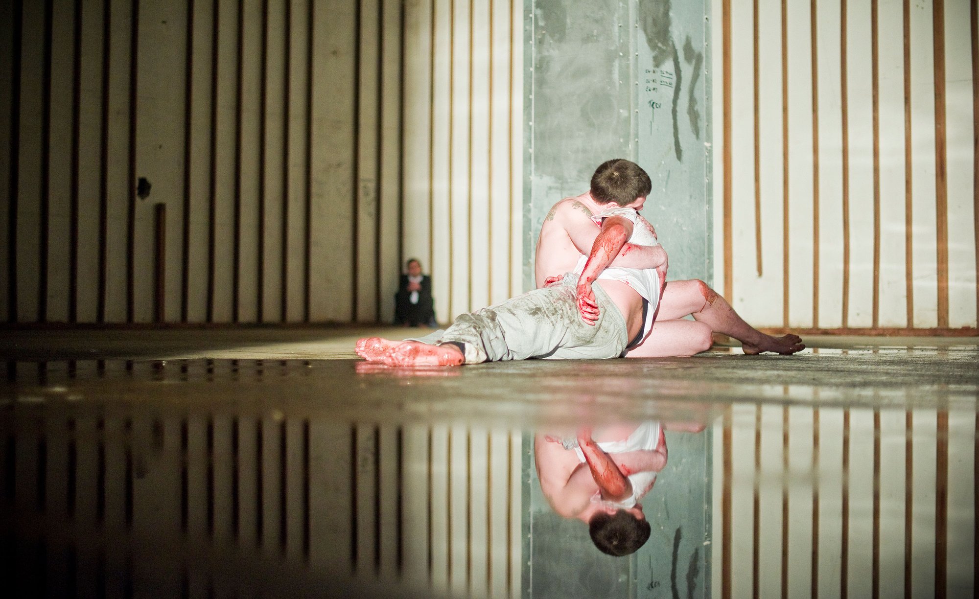 Image: A naked man sits cradling the lifeless bloody body of another man on the dank floor of a large industrial space, as an onlooker sits watching in the background during a tender subject, 2012. Photograph: Stephen White
