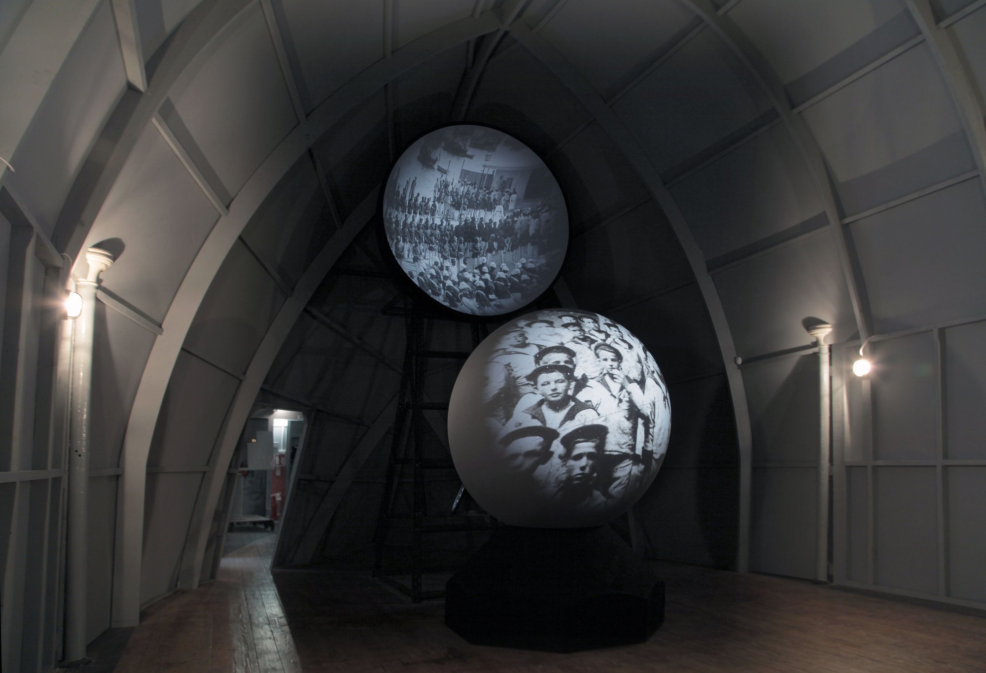 Image: Black and white images of sailors are projected onto two screens, concave and convex, as installed in The Tin Tabernacle, Kilburn, during Nowhere Less Now, 2012. Photograph: Lindsay Seers