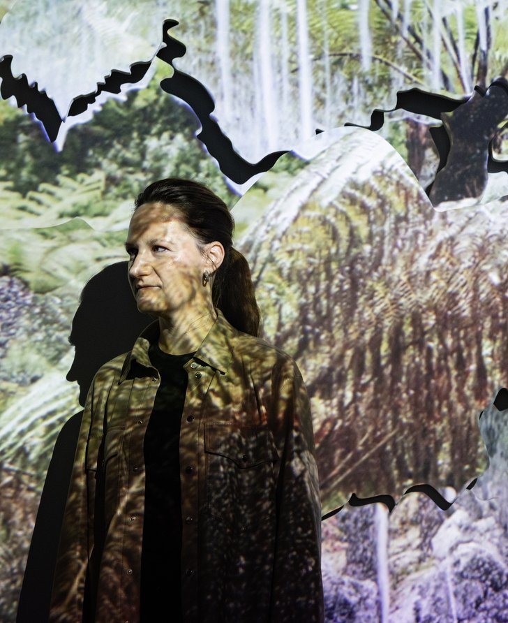 Noémie Goudal in front of her installation The Story of Fixity, with the video projection of foliage and drips over her.