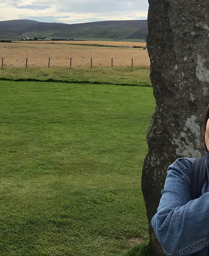 Dani Admiss in a denim jacket, leaning against a rugged stone wall with a green field as a backdrop.
