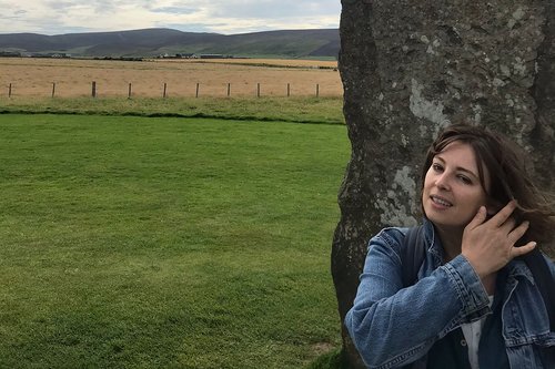 Dani Admiss in a denim jacket, leaning against a rugged stone wall with a green field as a backdrop.