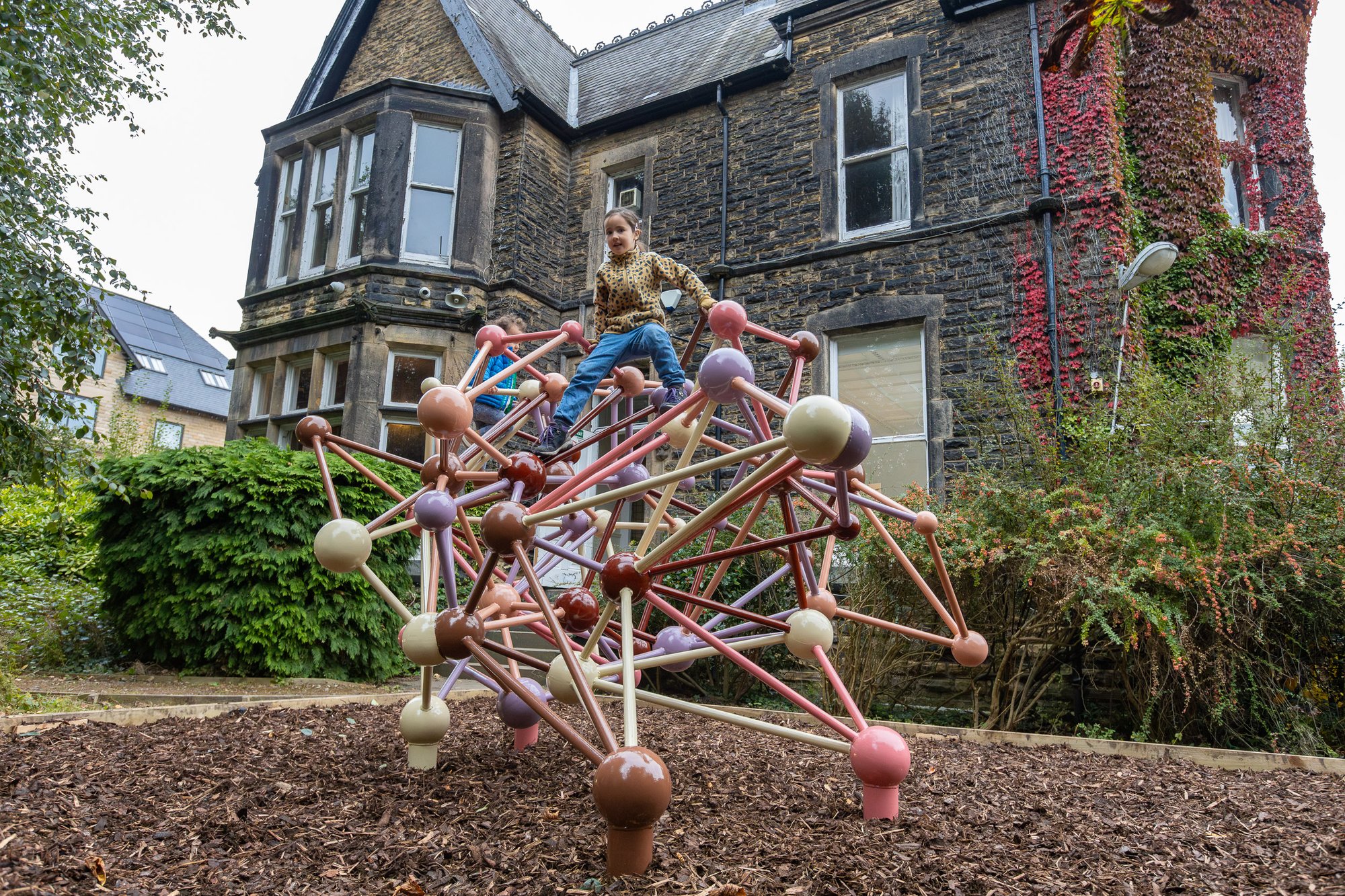 Two children playing on a 4D climbing frame that's been painted multiple colours, with a large Victorian house in the background.
