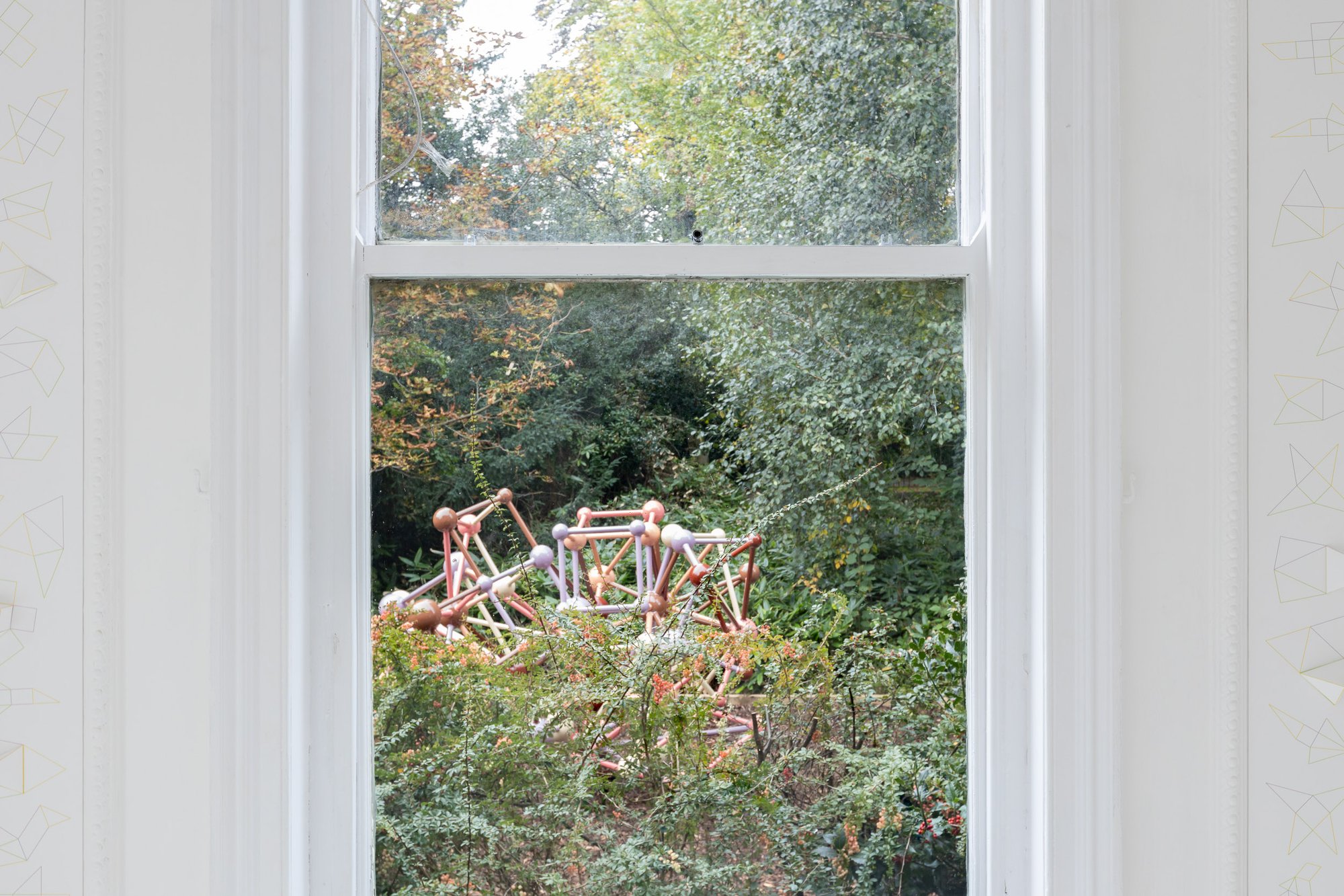 A colourful, geometric climbing frame peaking out of green foliage outside a window on a white wall.